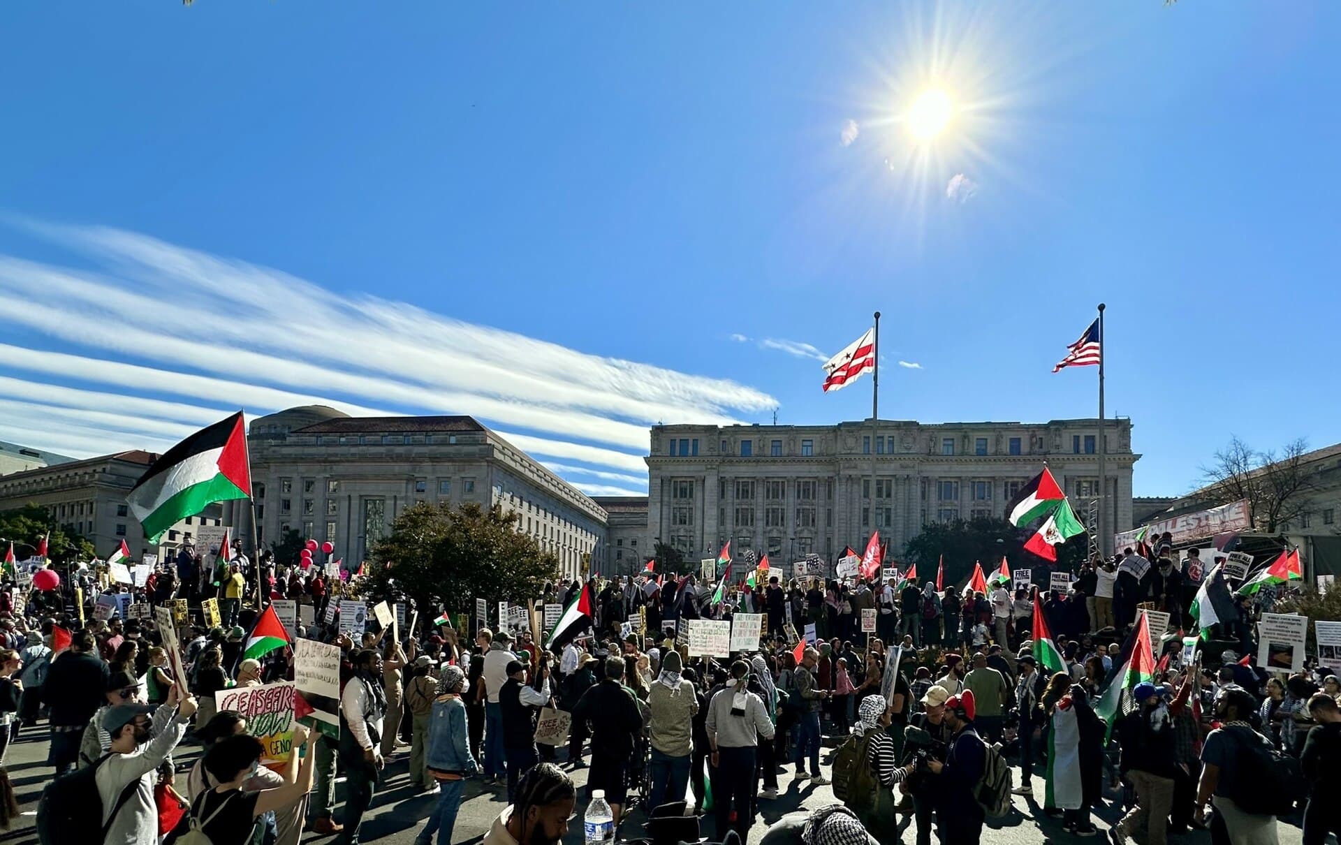 A dense crowd with Palestinian flags and hand-made signs in front of a stone government building, under a bright blue sky