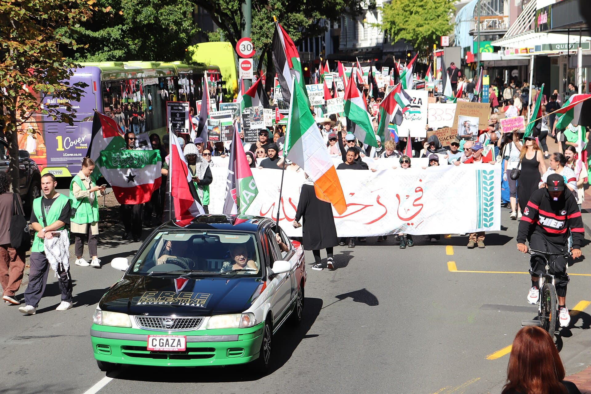 Marchers with flags and a long white banner walking down a city street behind a car decorated in Palestinian colours